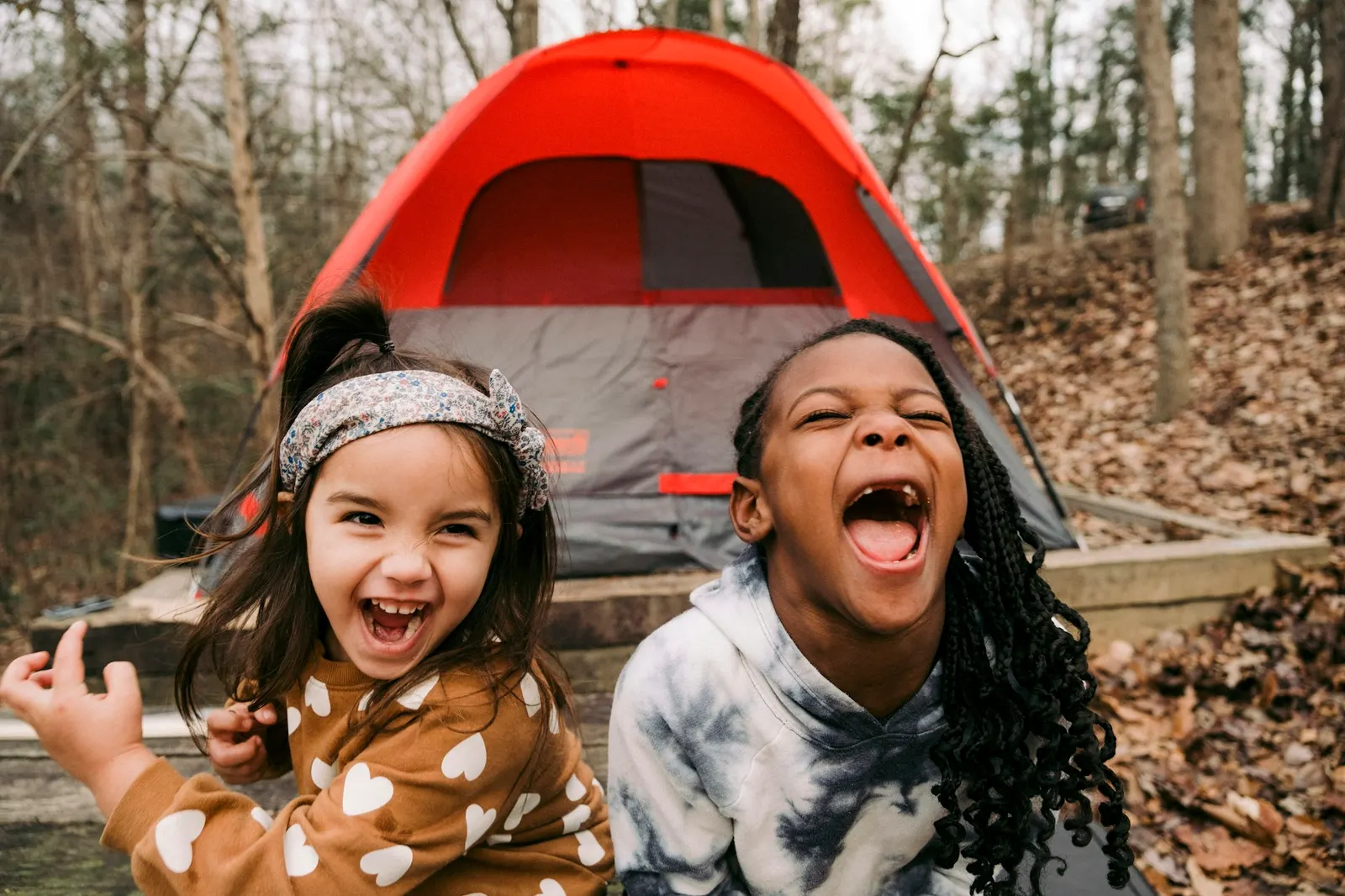 Two young girls laughing in front of a red tent at a forest campsite