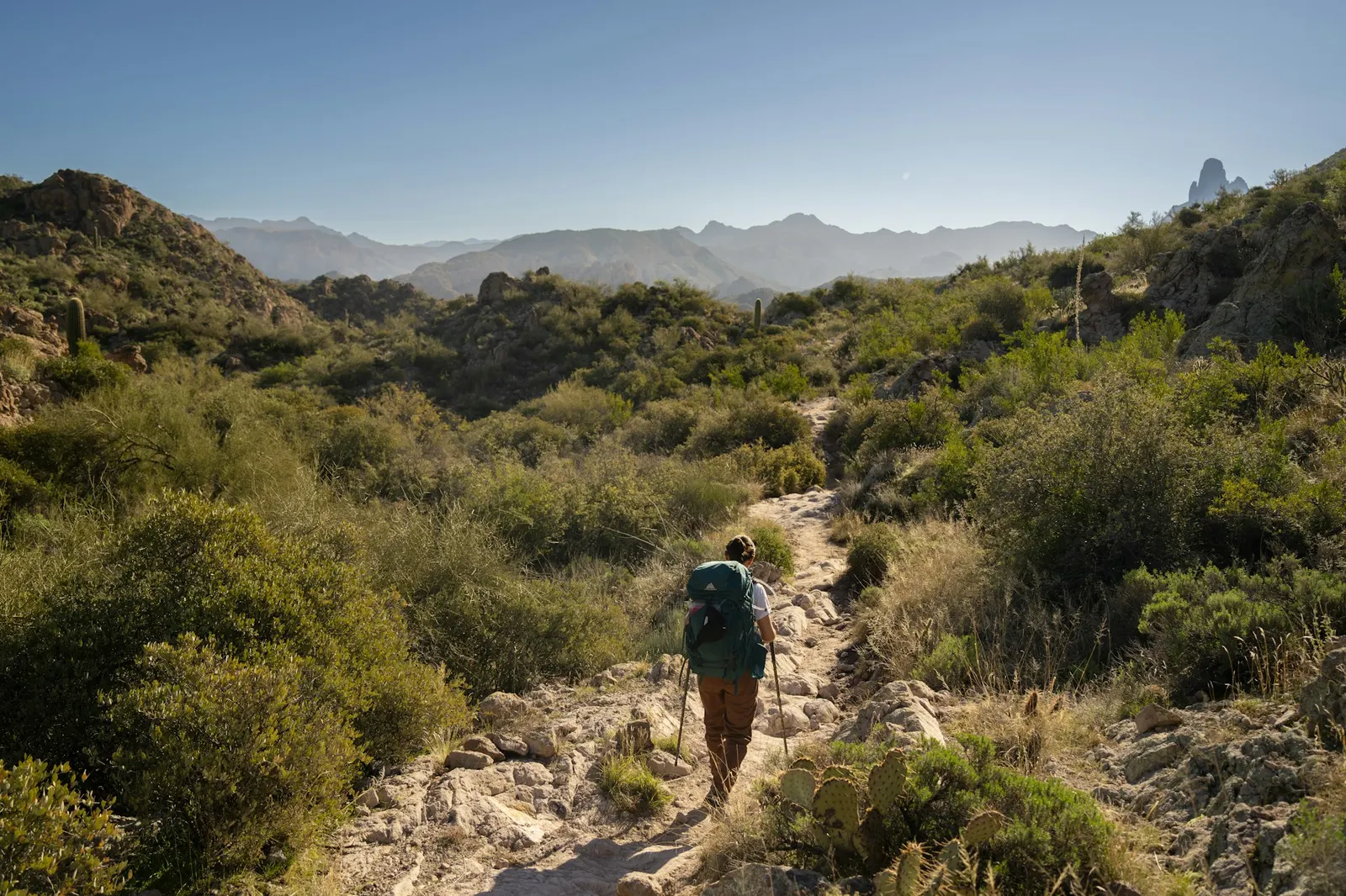 A hiker with a backpack walking on a rocky trail through green mountain shrubland