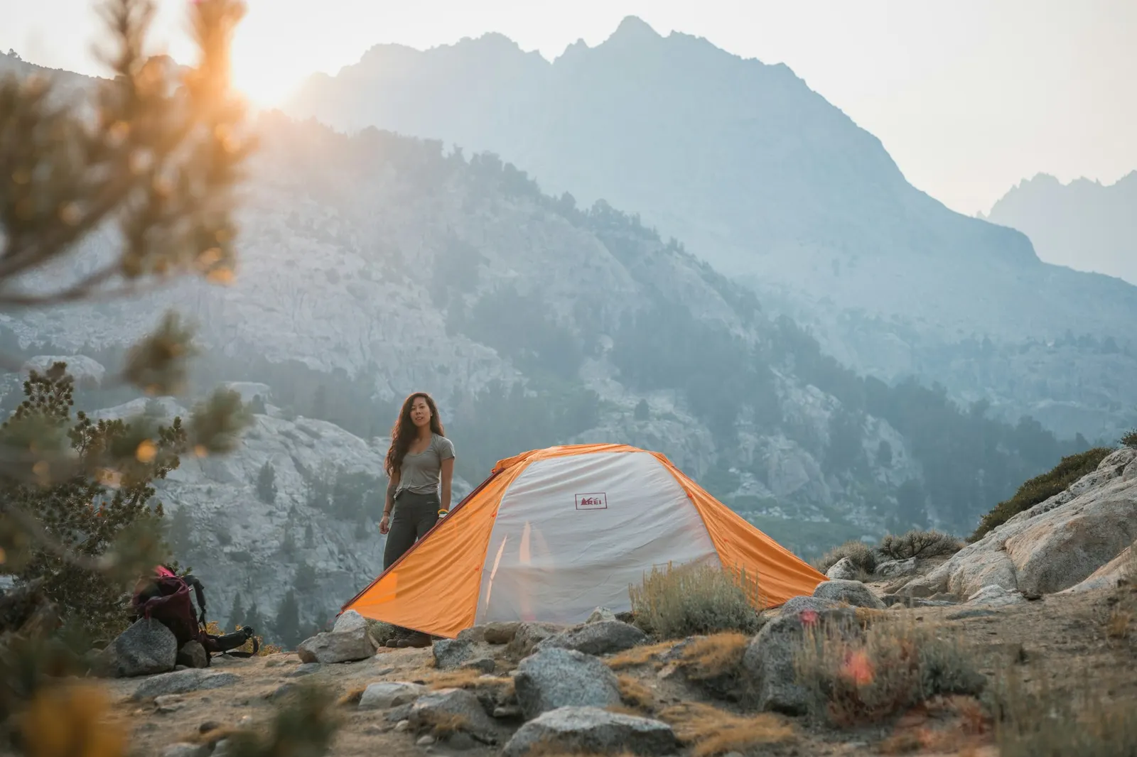 A hiker standing near an orange tent pitched among rocks on a mountainside at sunrise
